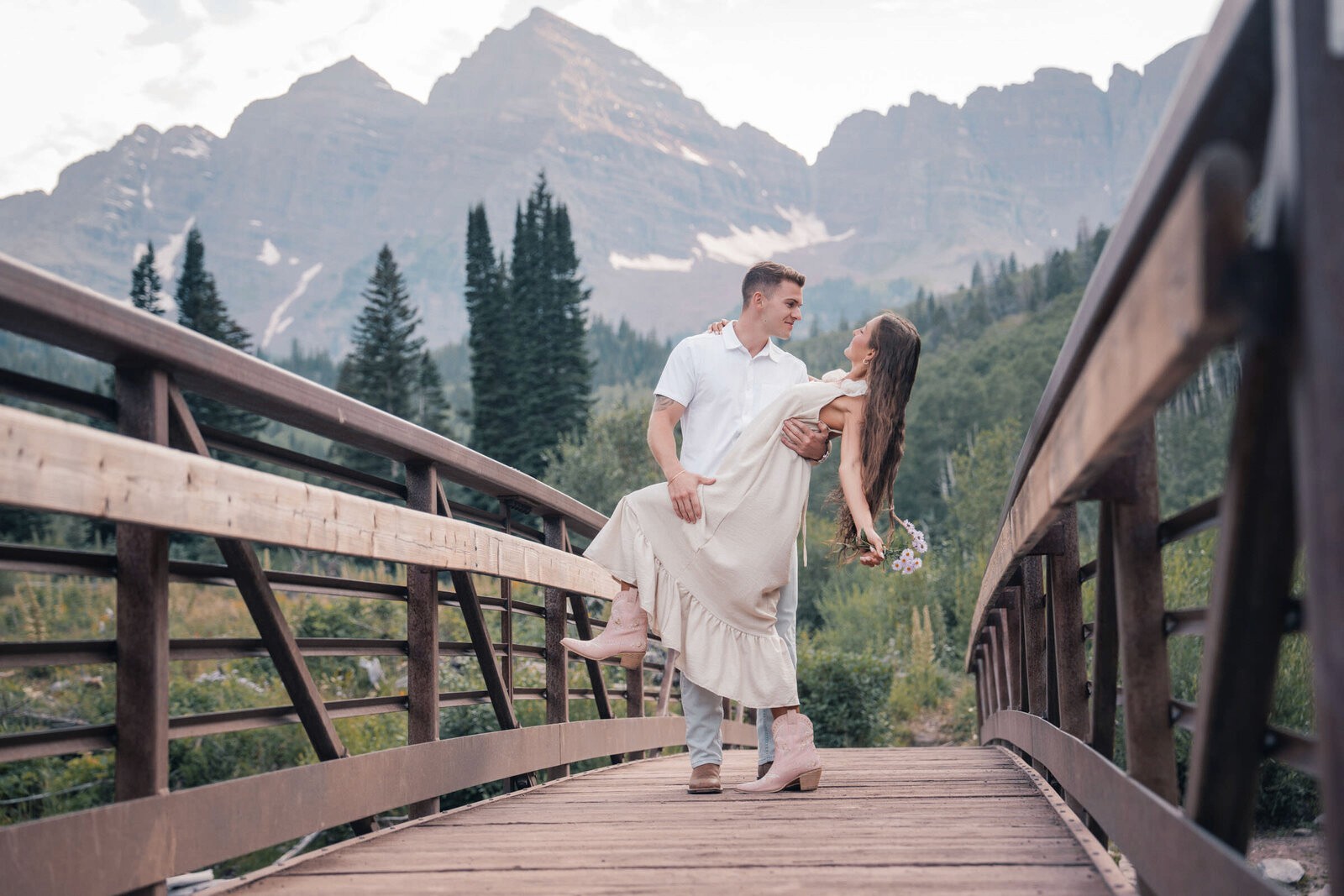 Man holding his partner on a wooden bridge, with evergreen trees and mountain peaks behind them.