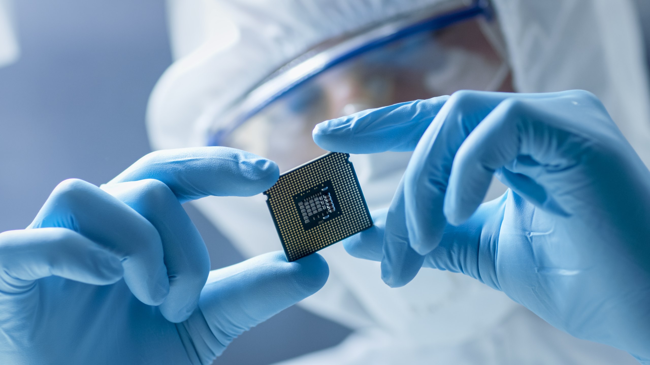 Technician in a cleanroom suit and blue gloves inspecting a microprocessor chip.
