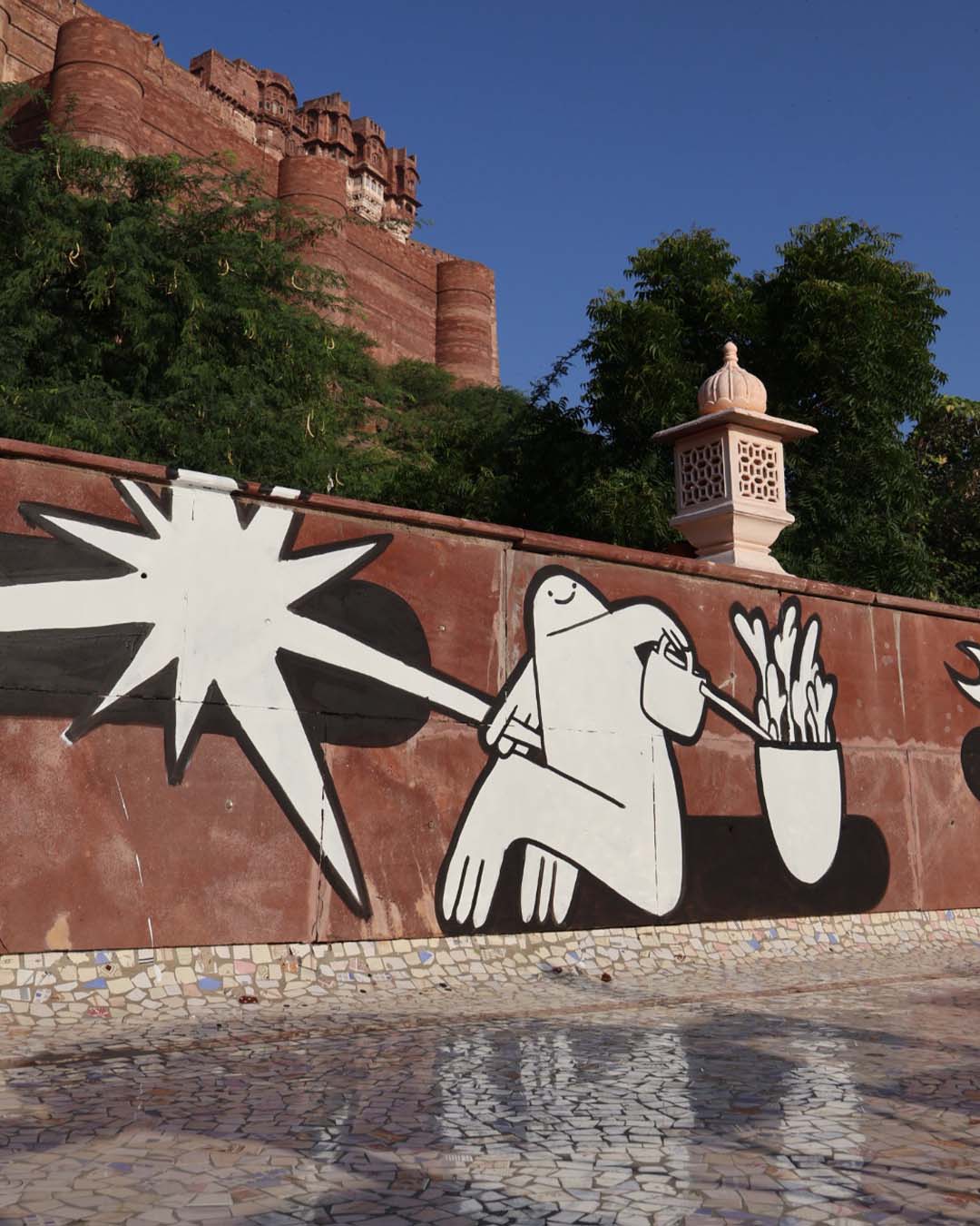 Mural painting of a joyful cartoon character watering a potted plant next to a star painted in black and white on a brown wall with oriental decorations visible and the Jodhpur fort in the background.