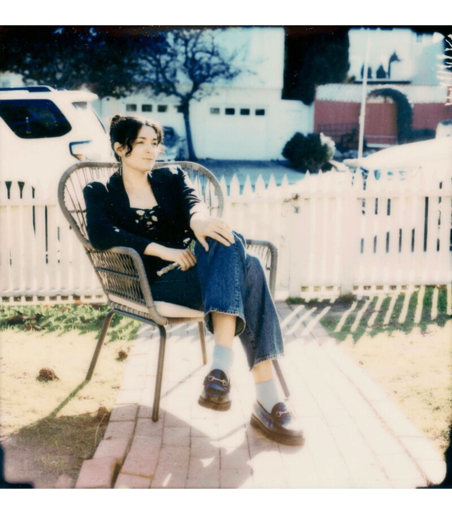 a polaroid of Rae Cohen with her hair up in a clip, wearing a blazer, jeans, and loafers in a wicker chair in a front yard in strong sunlight