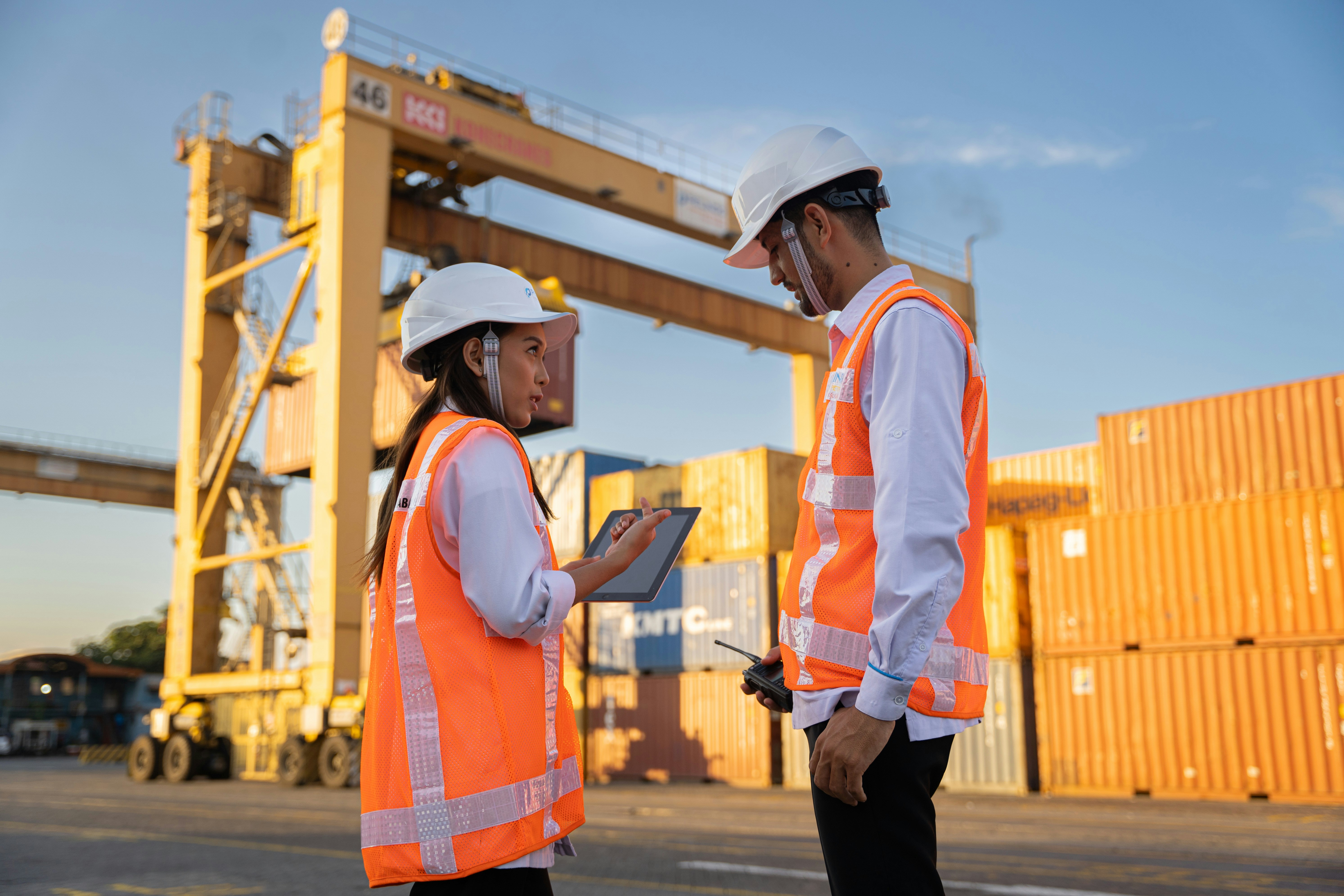 Two workers in hard hats discuss plans near shipping containers.