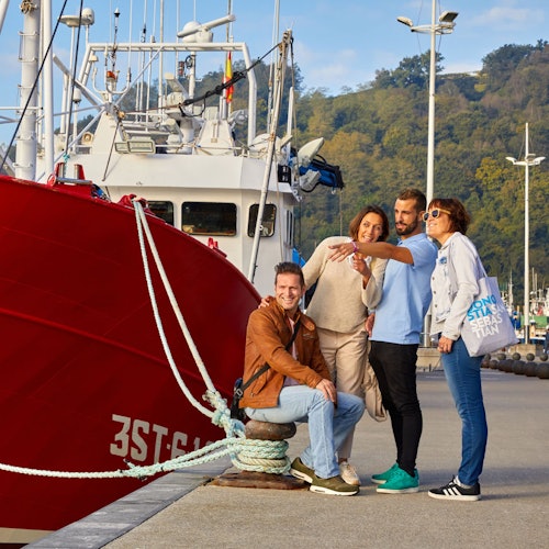 Four people smiling and posing for a photo by a docked red boat, with a wooded hillside in the background.