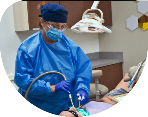 A dental professional in blue protective gear and mask performing a dental procedure on a patient under an overhead light