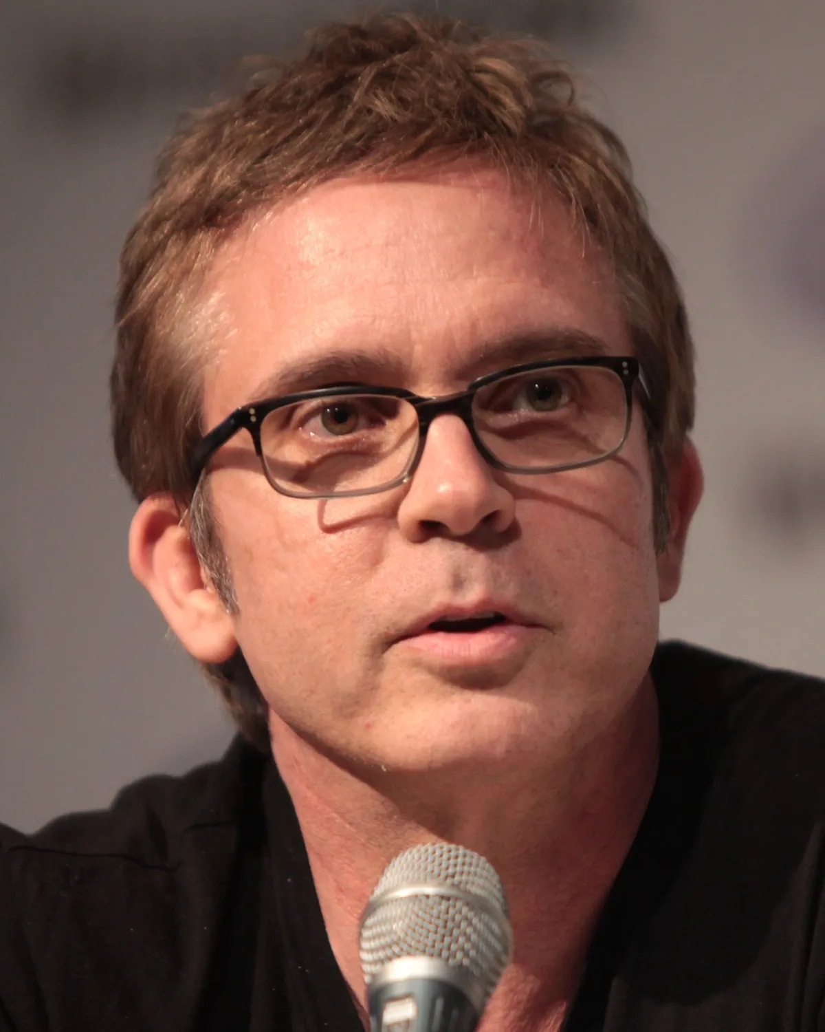 A man with short brown hair and glasses is speaking into a microphone, captured against a blurred indoor background.