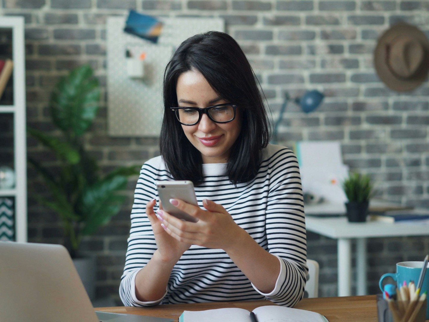 Smiling woman with curly hair and glasses using a smartphone in an office setting.