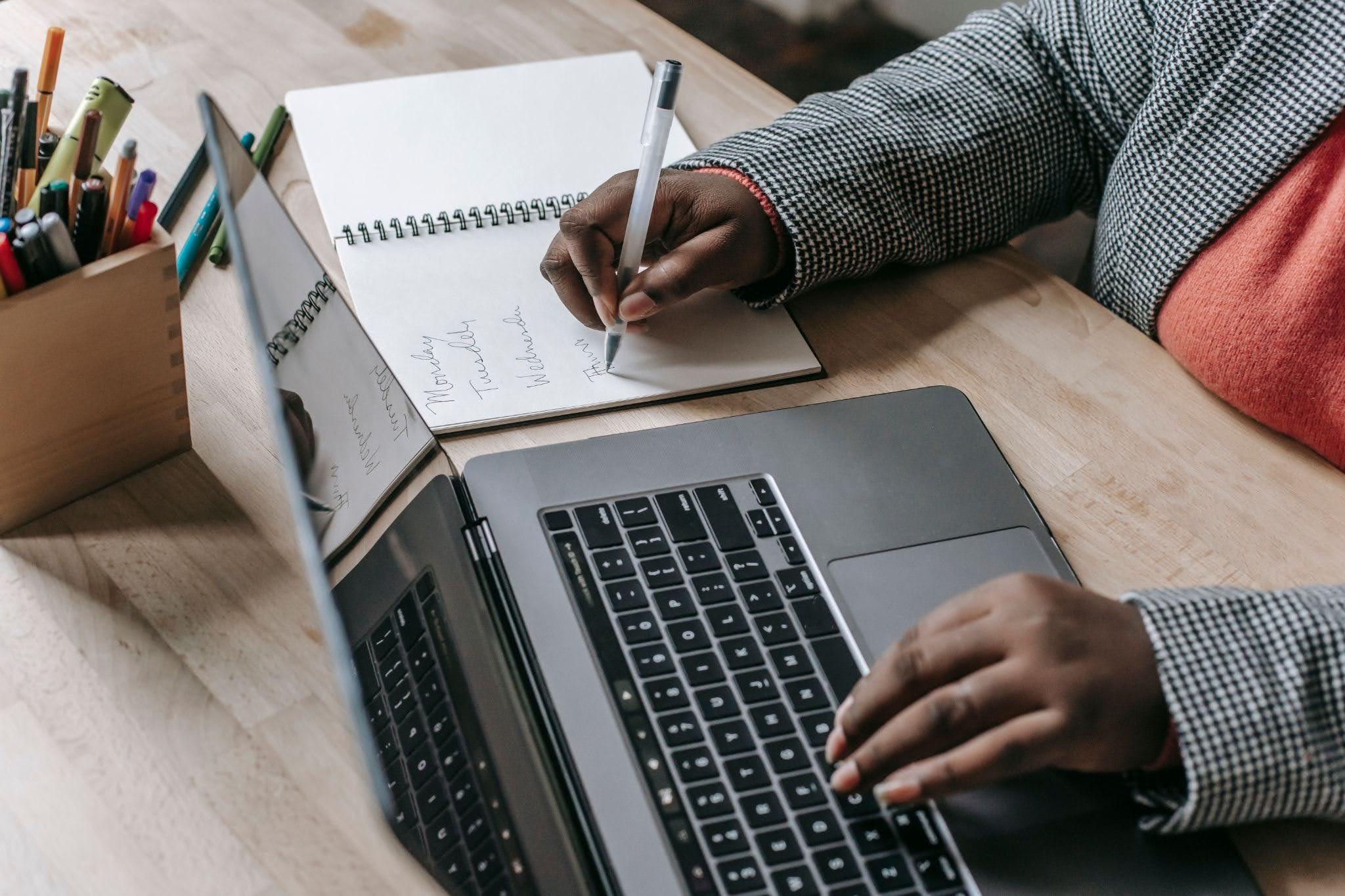 Individual worker writing notes on paper besides a laptop.