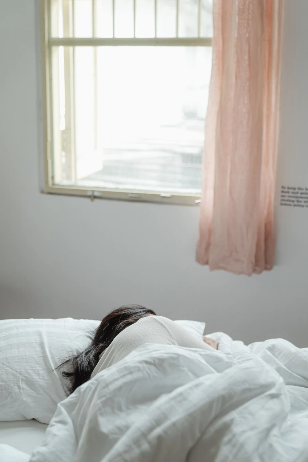 Person resting in bed beneath white bedding in a softly lit bedroom.