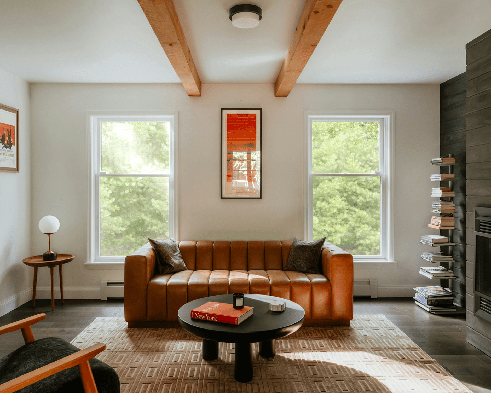 A modern living room features a brown leather sofa with black cushions, a round coffee table holding a book and candle, surrounded by earthy tones and decorated with wall art and stack of books by the fireplace, illuminated by natural light from large windows.
