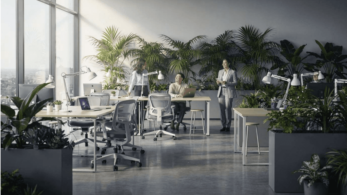 Modernes, helles Büro mit vielen Pflanzen: Team arbeitet gemeinsam an Schreibtischen, spricht miteinander und nutzt Laptops in einer entspannten, gesunden Arbeitsumgebung.