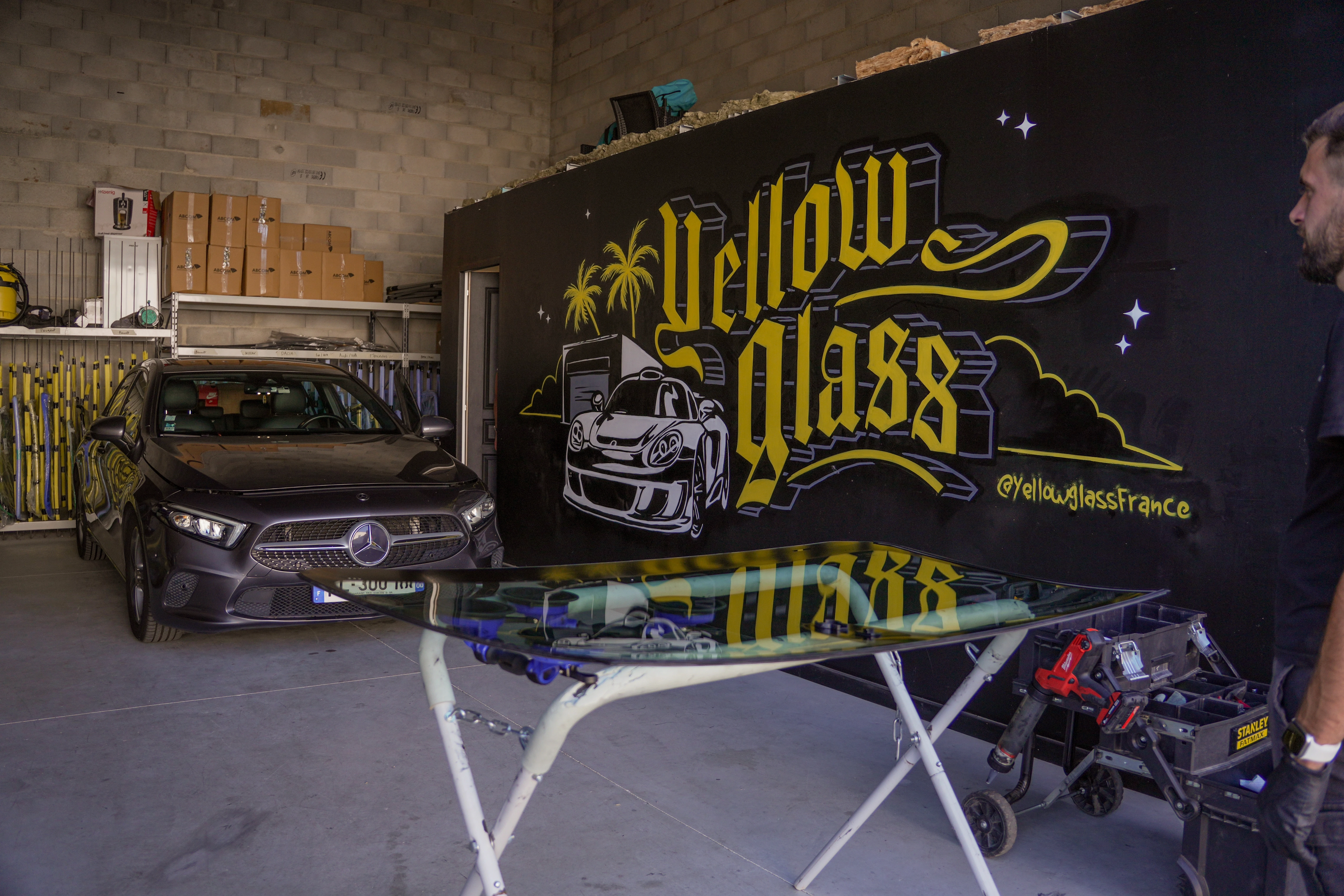 Une agence de remplacement de pare-brise avec une voiture Mercedes noire devant un mur avec un logo "Yellow Glass". Réparation automobile professionnelle.