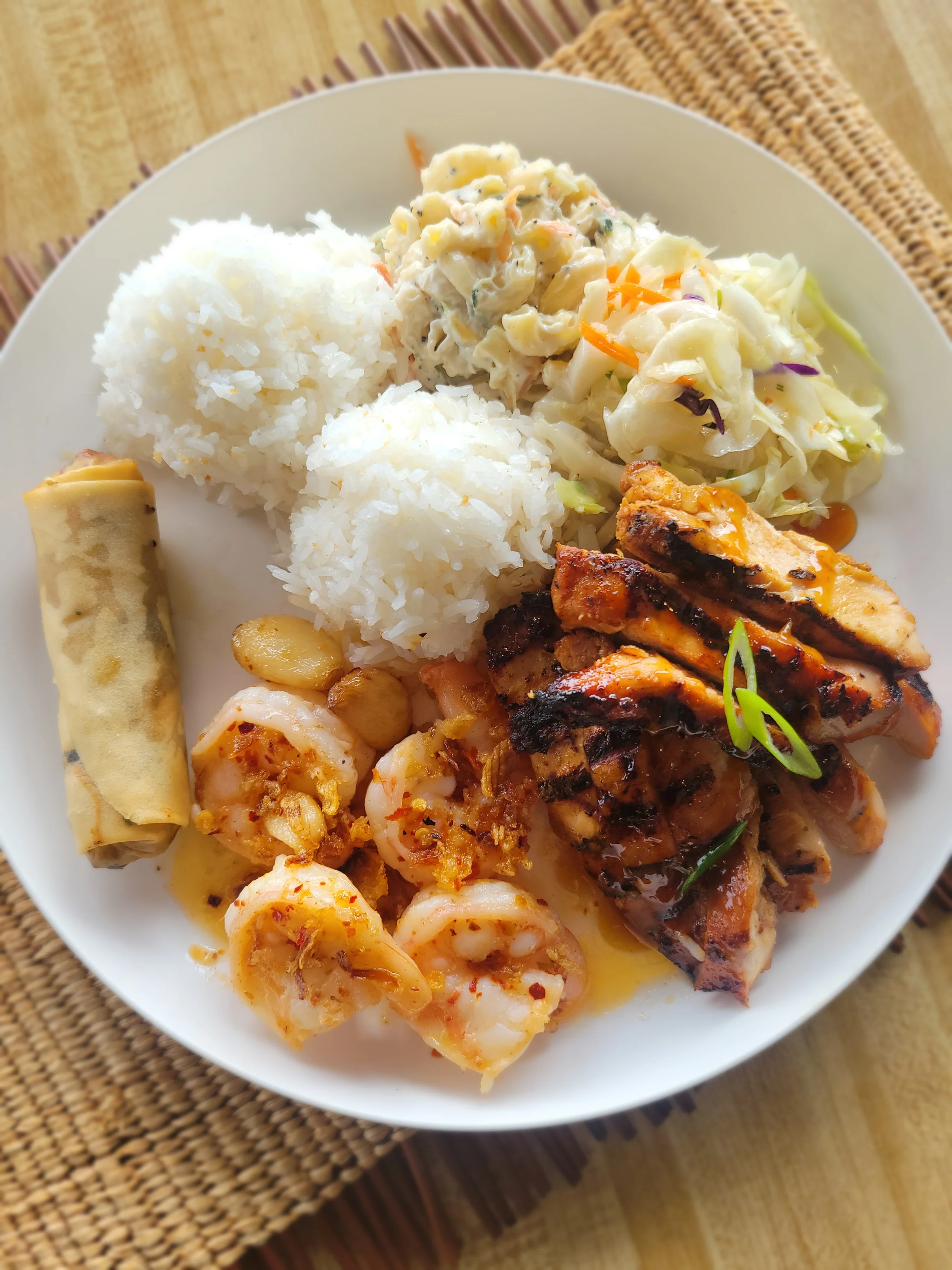 A large white plate featuring sliced grilled Huli Huli chicken and several pieces of buttery garlic shrimp, served with a spring roll, white rice, macaroni salad, and slaw.