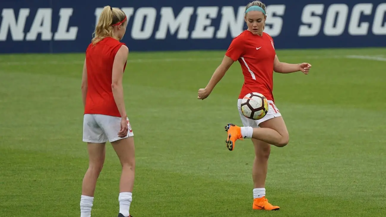 Two footballers in red shirts are training on a green football pitch. One player juggles the ball while the other watches attentively. A sign reading ‘WOMENS SOCCER’ can be seen in the background. The picture illustrates the rise and dynamism of women's football.