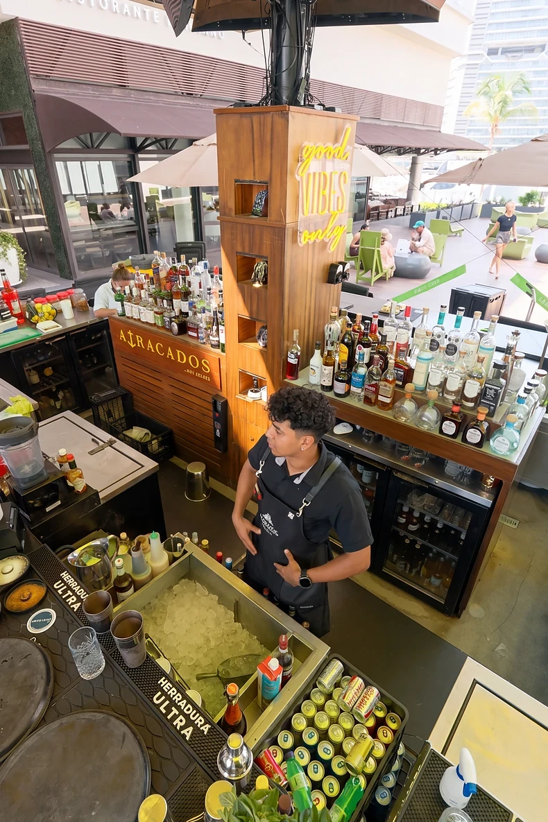 Vista desde arriba de la barra de Atracados con un bartender, botellas y el neón "good vibes only"