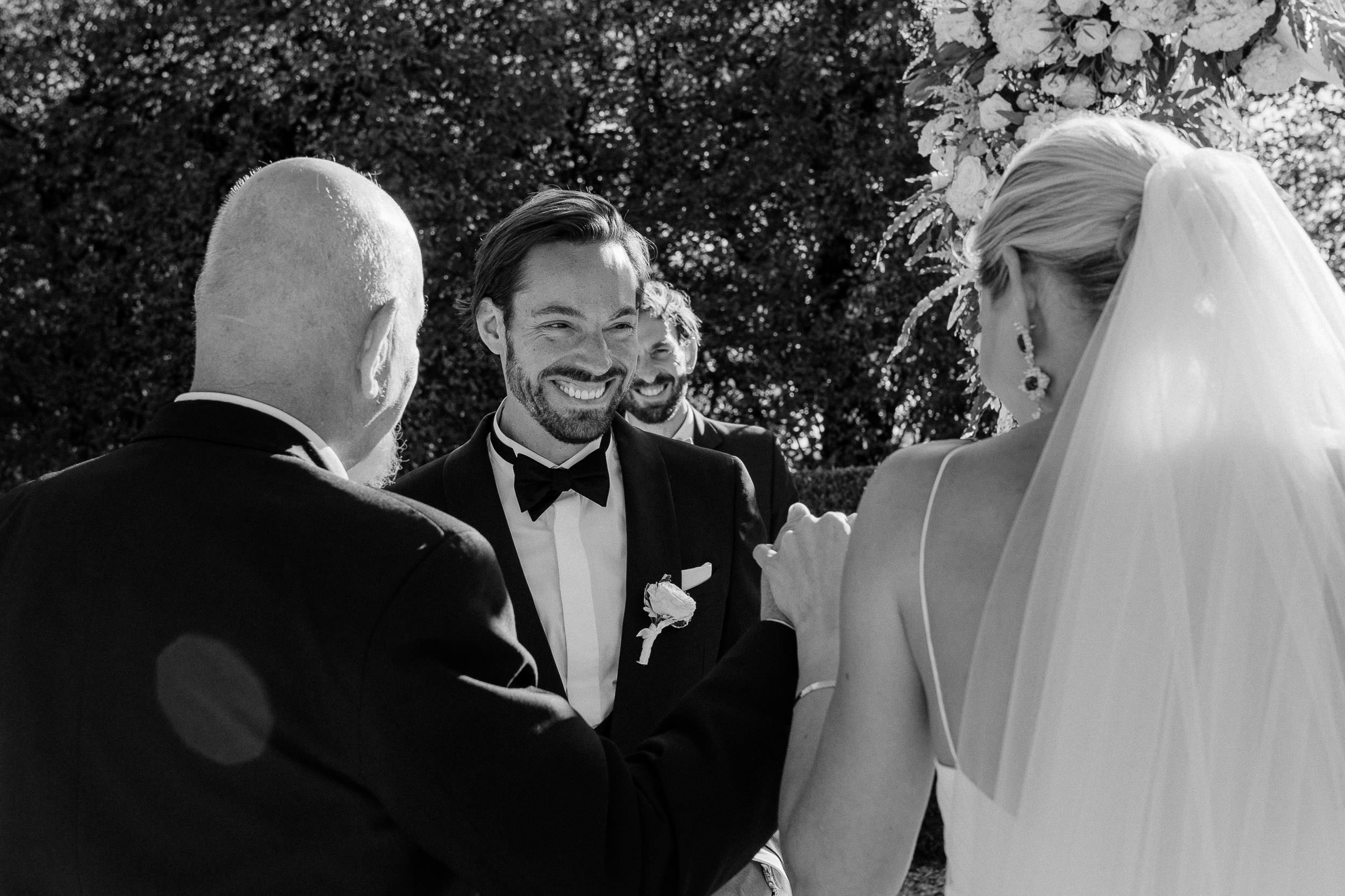 A groom in a tuxedo smiles at a bride in a veil as they exchange vows with an officiant in a black suit.