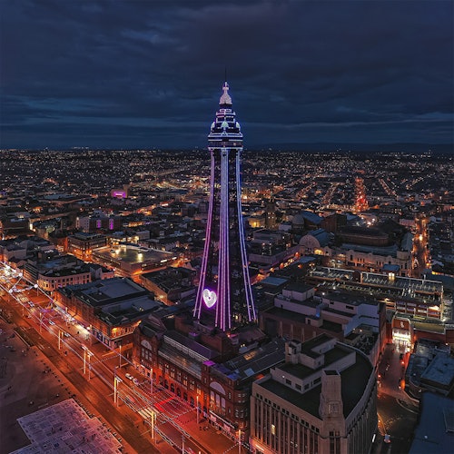 A night view of a city featuring a tall, illuminated tower with colorful lights amidst surrounding buildings and streets.