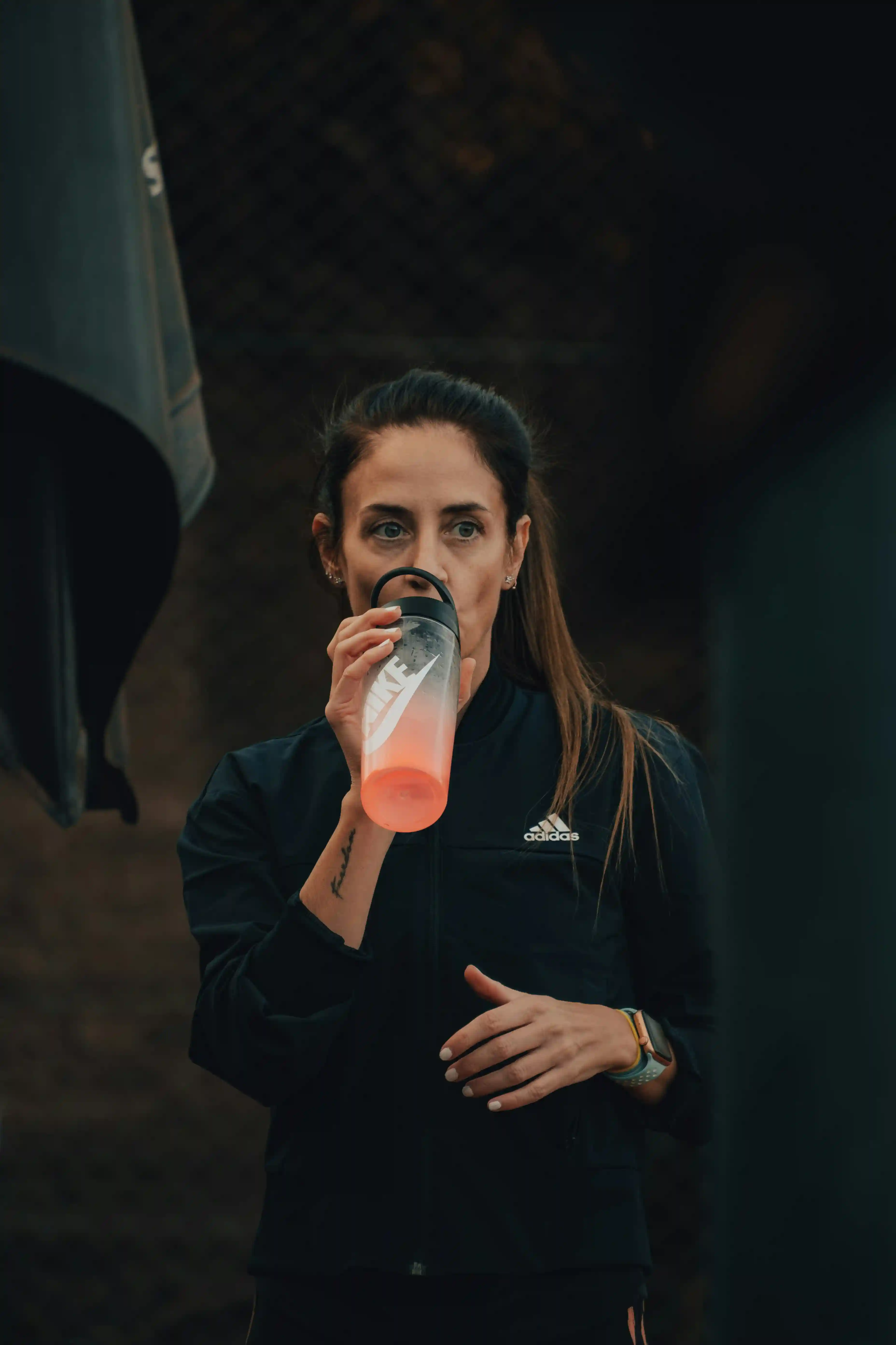 Female athlete drinking from a shaker bottle outdoors, wearing a black training jacket.