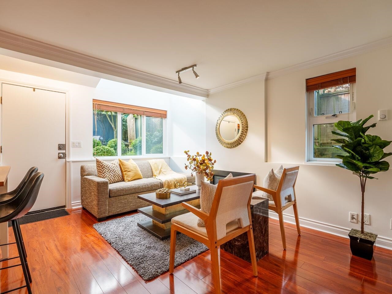 Cozy living room with hardwood floors, a couch, dining table, and sunlight from large windows. Green plant in corner.