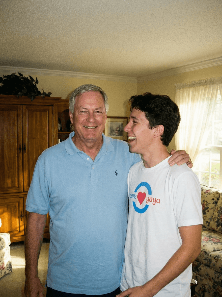 An older adult man in blue polo stands smiling with arms around young man in CareYaya T-shirt in a living room