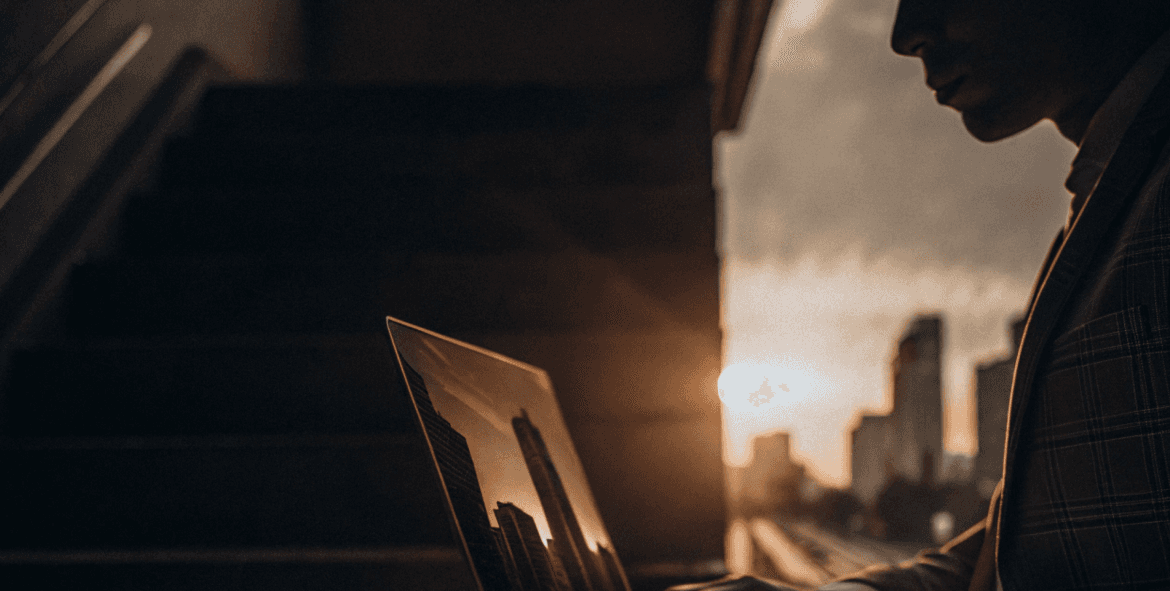Person working on a laptop with a city background during sunset, with a person in a suit.