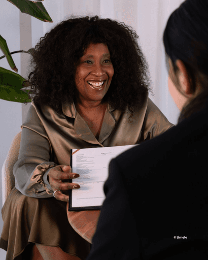 Een trouwambtenaar met krullend haar glimlacht terwijl ze een document aan een ander overhandigt en in gesprek is.