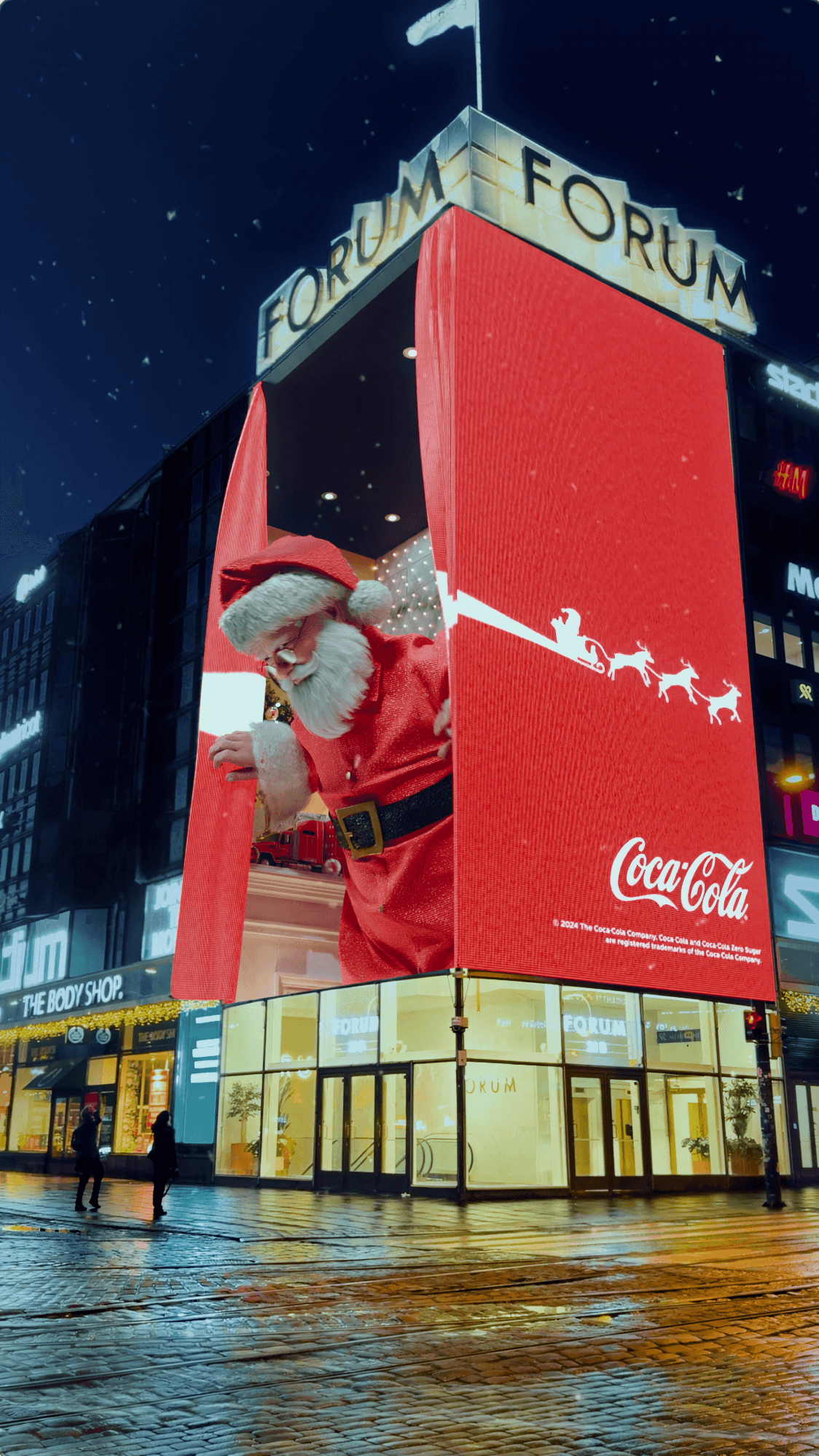 Giant Santa on a building screen reaches for a glowing Coca-Cola Christmas truck on a wet city street at night.