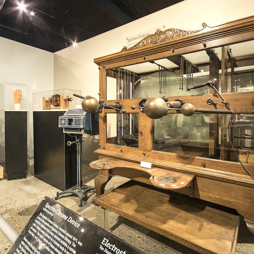 一個展示復古醫療設備的博物館展覽，包括一台帶金屬球的木製電療機和各種儀器的展示。