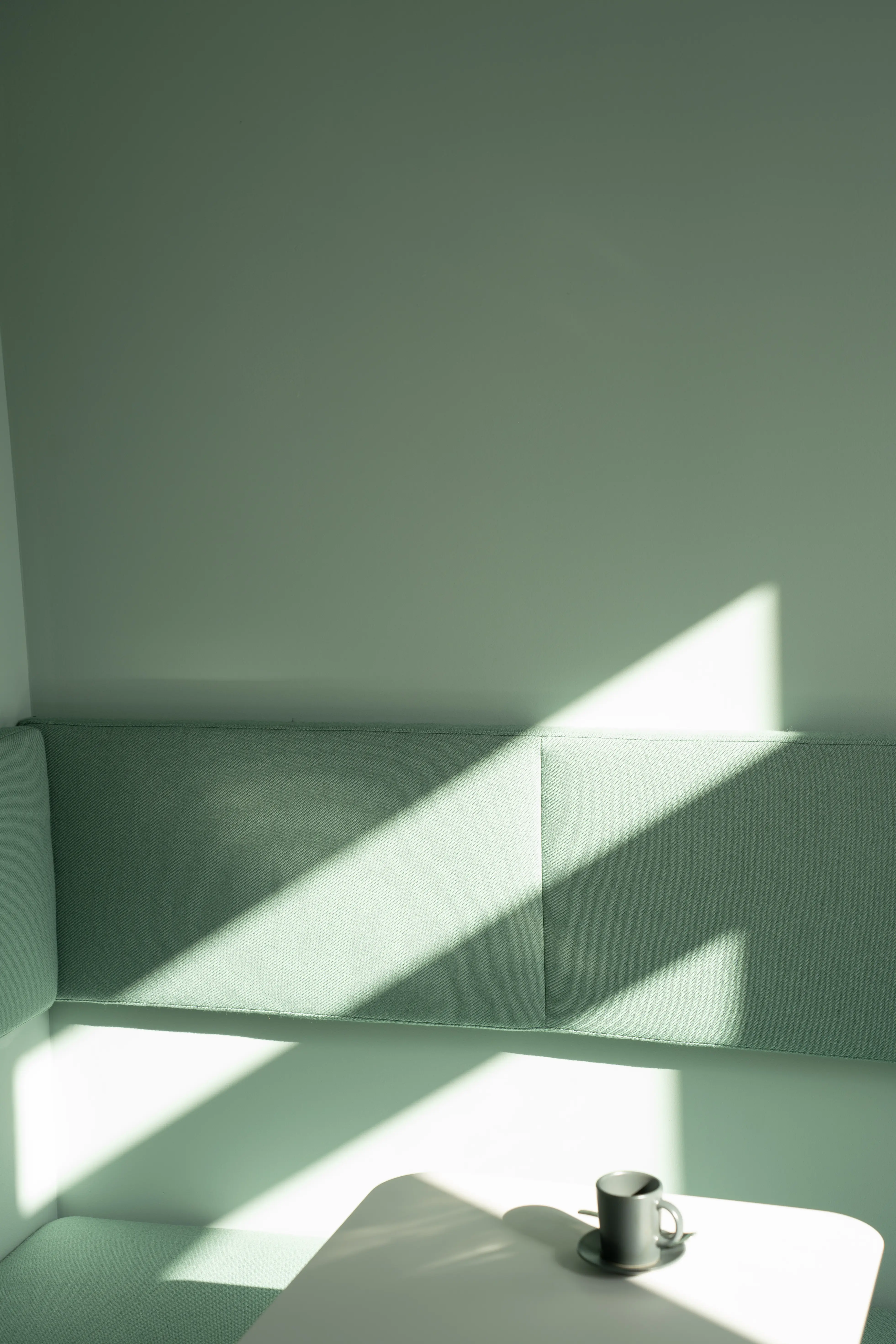Sunlight falling across a mint-green upholstered bench niche at AI START-UP MUNICH with a matching green wall and a white table holding an espresso cup.