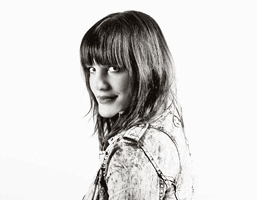 A black and white portrait of a young woman with shoulder-length dark hair and bangs. She is looking over her shoulder towards the camera. She is wearing a denim jacket with a visible collar and straps. The background is plain white.