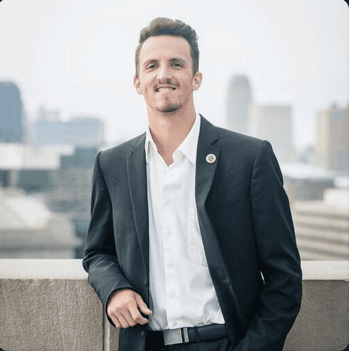 A person with curly hair and glasses smiles at the camera, seated in a cozy workspace with warm lighting.