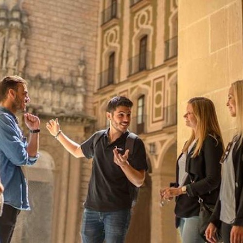 Three people listening to a fourth person speak, standing in an ornate courtyard with detailed building facades.