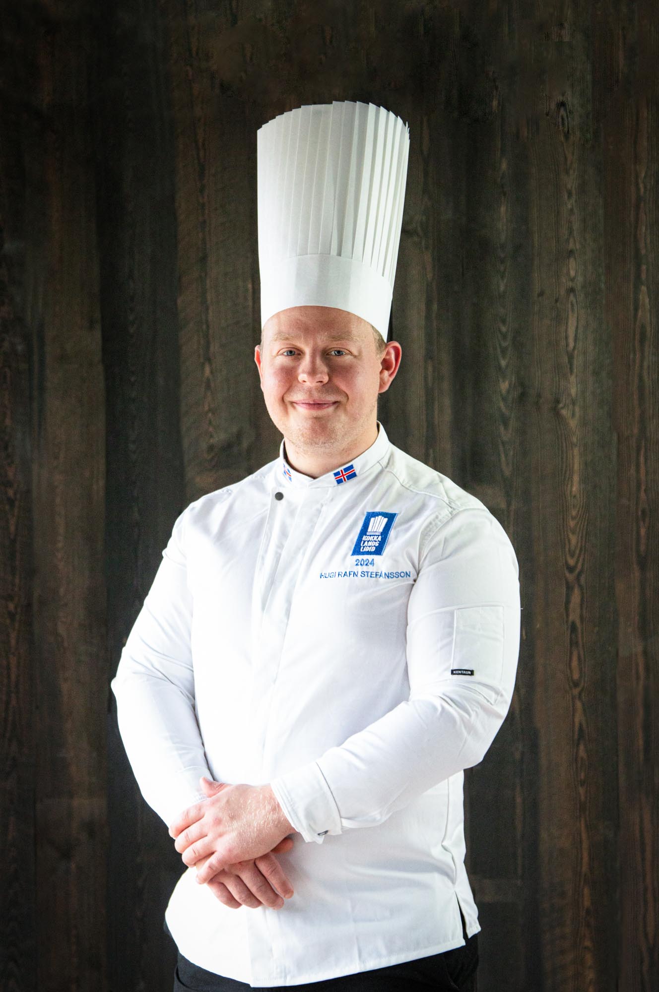 Chef Hugi Rafn Stefánsson smiles in a portrait, wearing a tall white chef's hat and the Icelandic National Culinary Team's white jacket, against a dark wood-panel background.