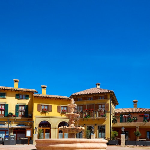Sunny courtyard with a central fountain, surrounded by colorful, Mediterranean-style buildings under a clear blue sky.