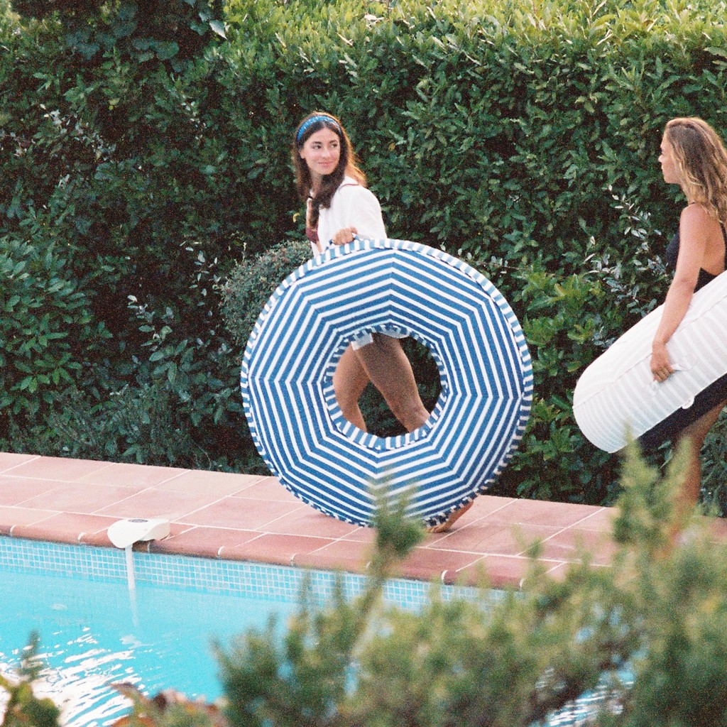 Two women with a luxury blue-and-white pool float poolside at a Mediterranean garden with terracotta tiles