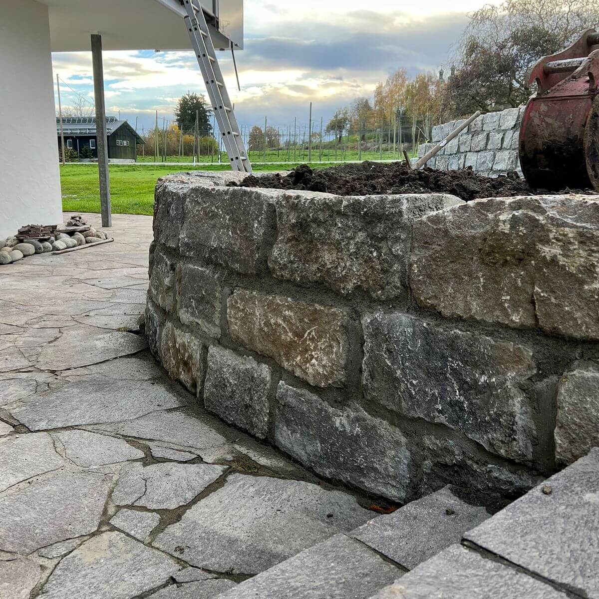 Ansicht einer fertiggestellten, stabilen Mauer aus dunklen Natursteinen zur Hangsicherung in einem Garten, Gartenbau Klingenstein Berg.