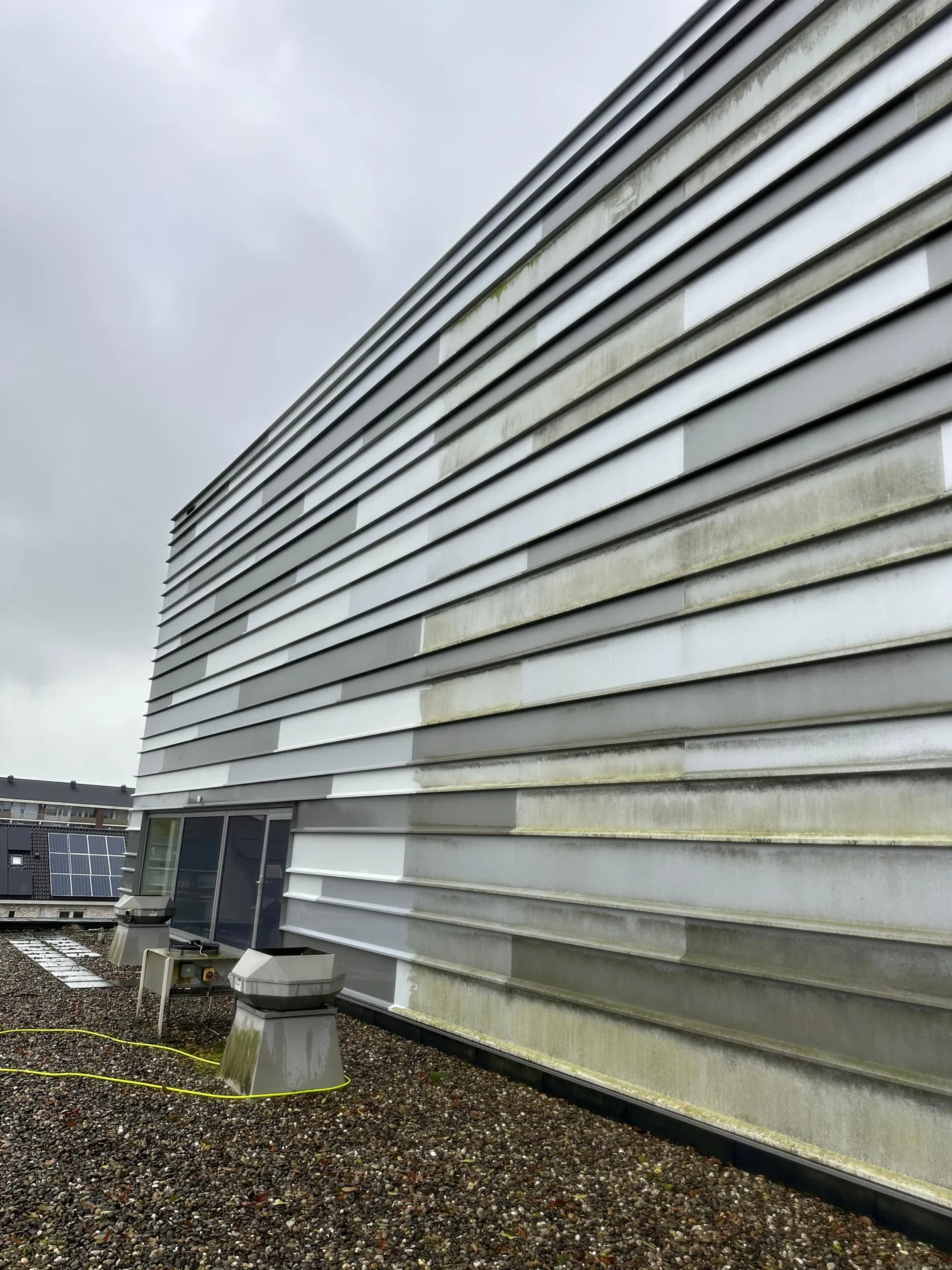 Hoge gevel met horizontale grijze lamellen gezien vanaf een grinddak: het voorste deel is schoon lichtgrijs, verderop zitten groene aanslag en donkere regenstrepen op de panelen