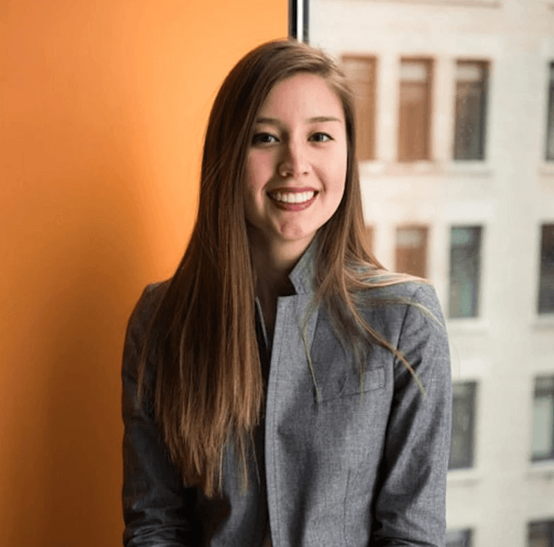 Young woman with long brown hair smiles while seated in a gray suit jacket against an orange wall and large window. Urban building in background.