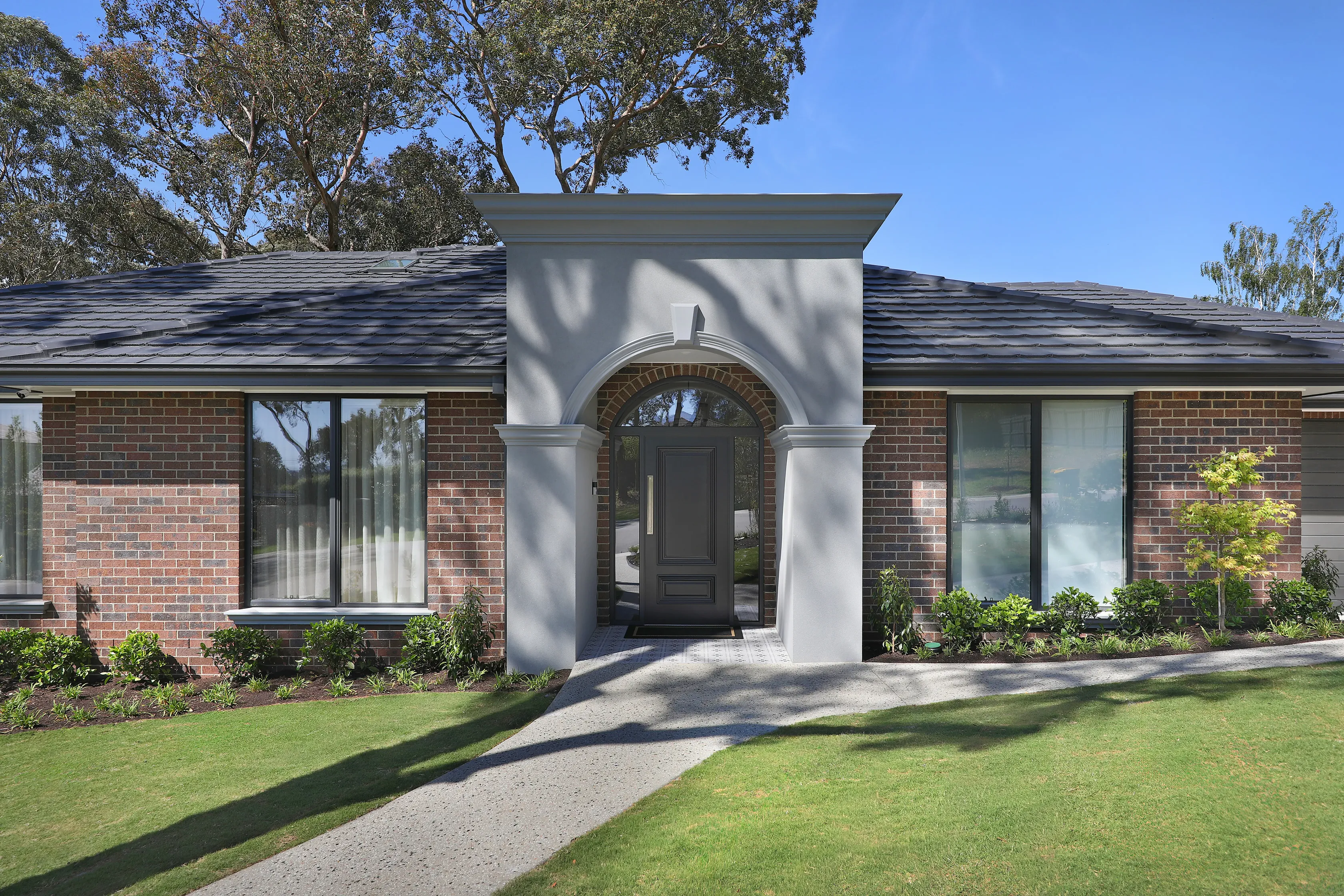 Entry of the Diamond Creek home, a single-storey blend-brick residence with a rendered arched portico, dark grey tiled roof, black panelled door and an aggregate path through a lawn beneath gum trees