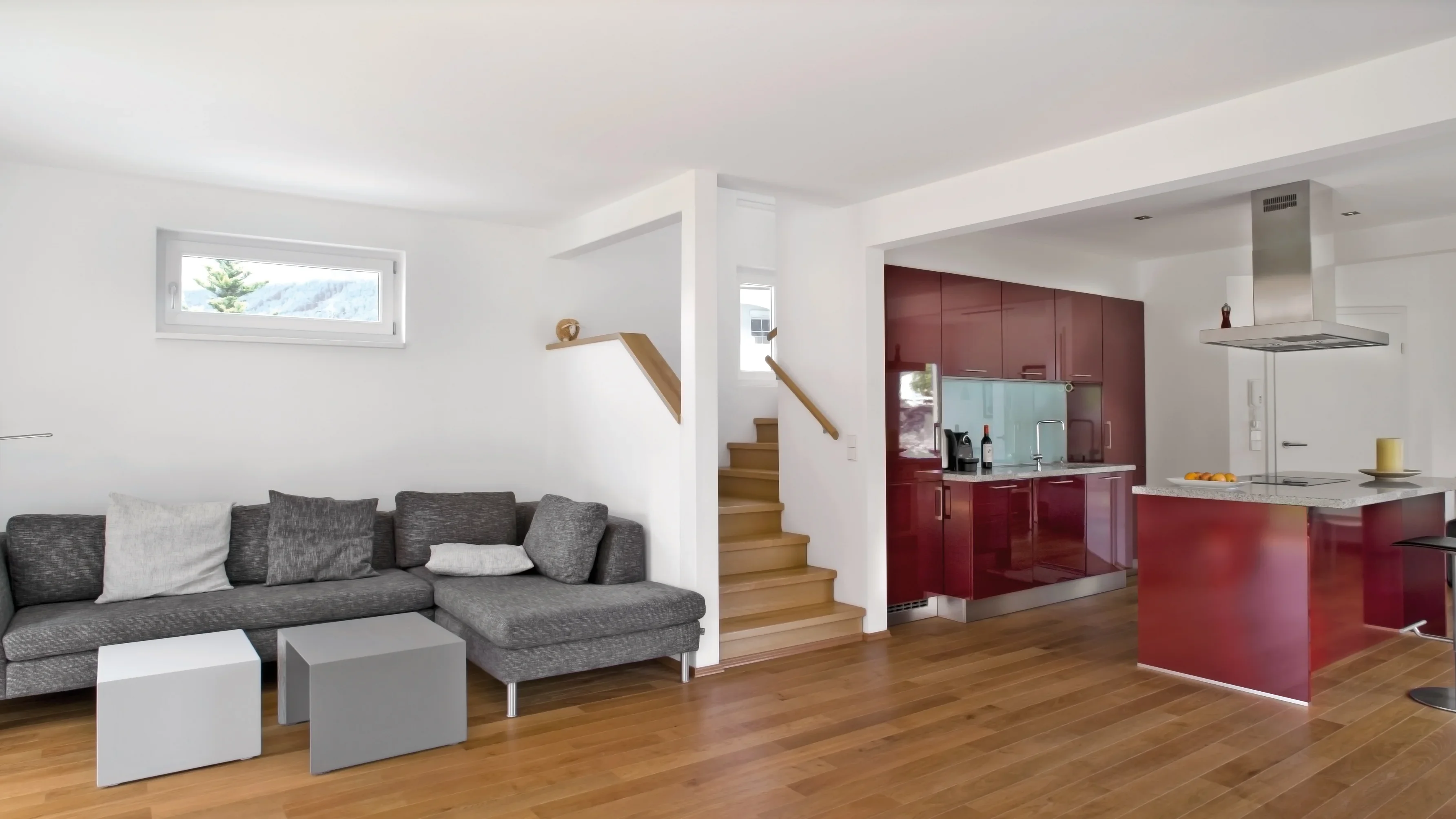 Moderne Wohnküche mit roter Küchenzeile und Kochinsel, daneben ein Wohnbereich mit grauem Ecksofa und Holzboden.