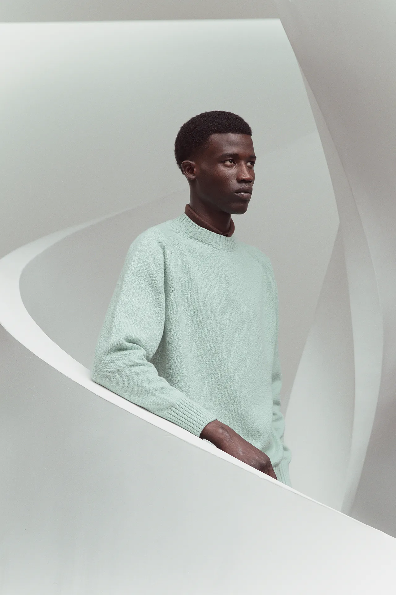 Aida Barni SS26 collection men's mint green crewneck knit sweater worn over a brown shirt by a model standing on a white spiral staircase.
