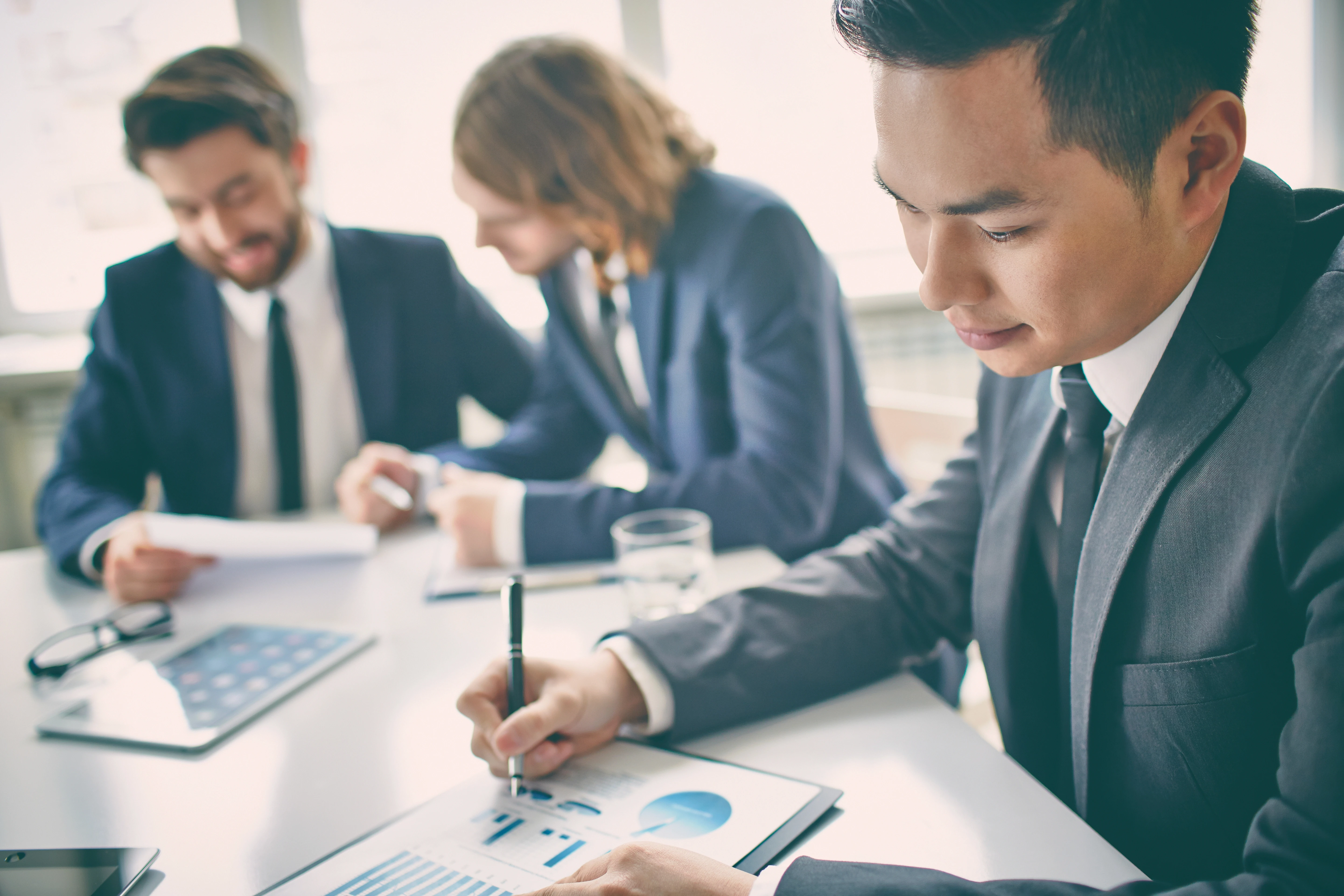 Three business professionals in suits review financial documents at a meeting table, with one person writing notes on a chart while others discuss paperwork in the background.