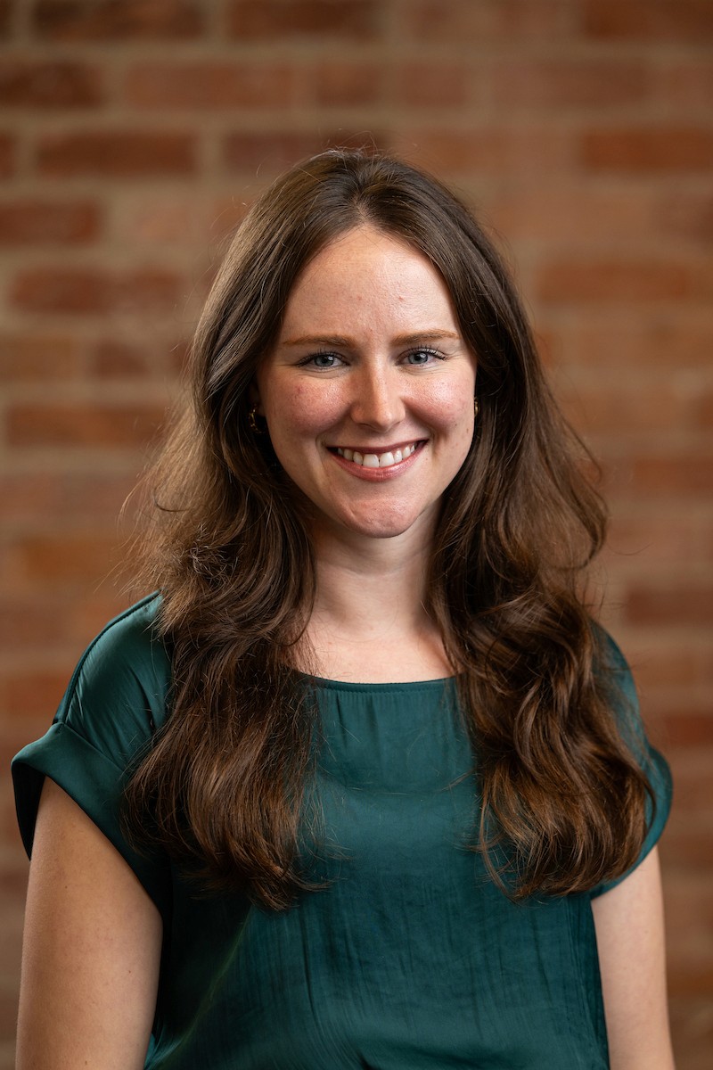 A woman with long brown hair smiles in front of a brick wall, wearing a green shirt.