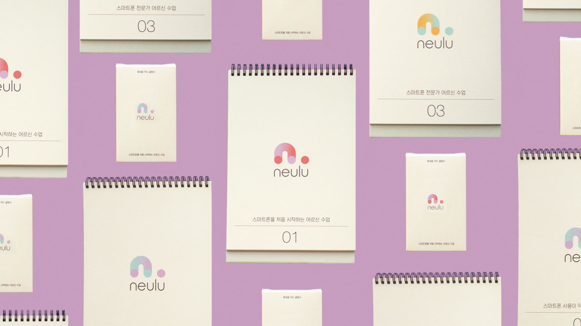 Multiple copies of a Neulu branded spiral-bound notebook are arranged on a light purple background. Each notebook features the brand’s logo: a stylized rainbow-colored circle with a curved line above it, and the word “neulu” written below. The notebooks are numbered “01” and “03” on the front and back, respectively, with Korean text printed beneath. The notebooks are displayed at various angles and orientations.