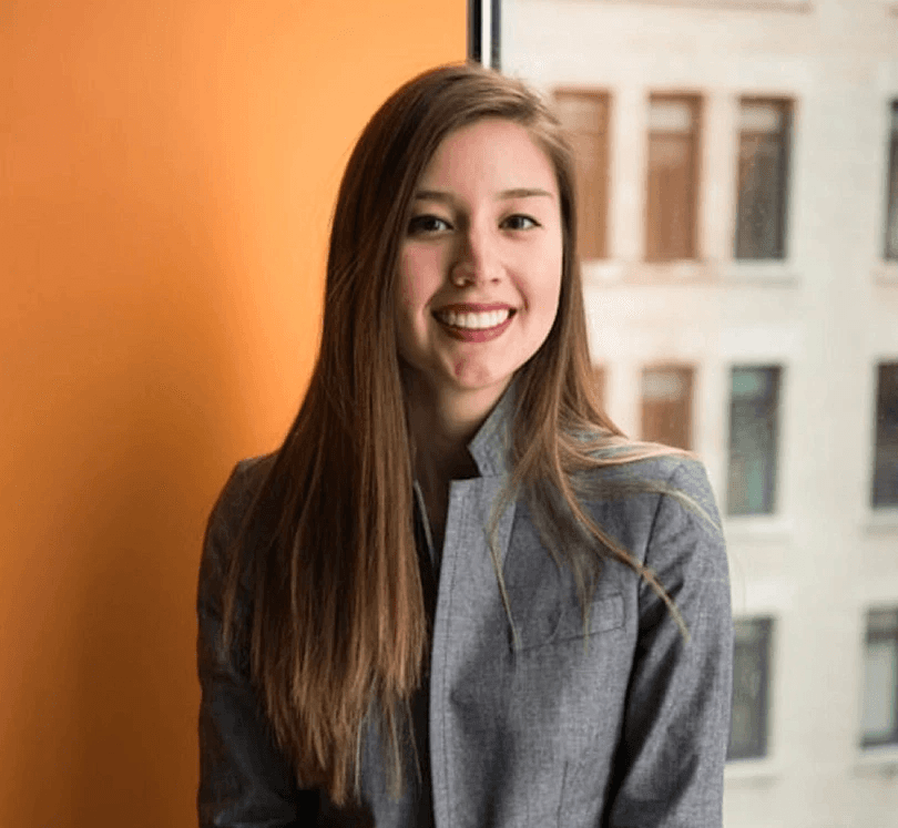 Young woman with long brown hair smiles while seated in a gray suit jacket against an orange wall and large window. Urban building in background.