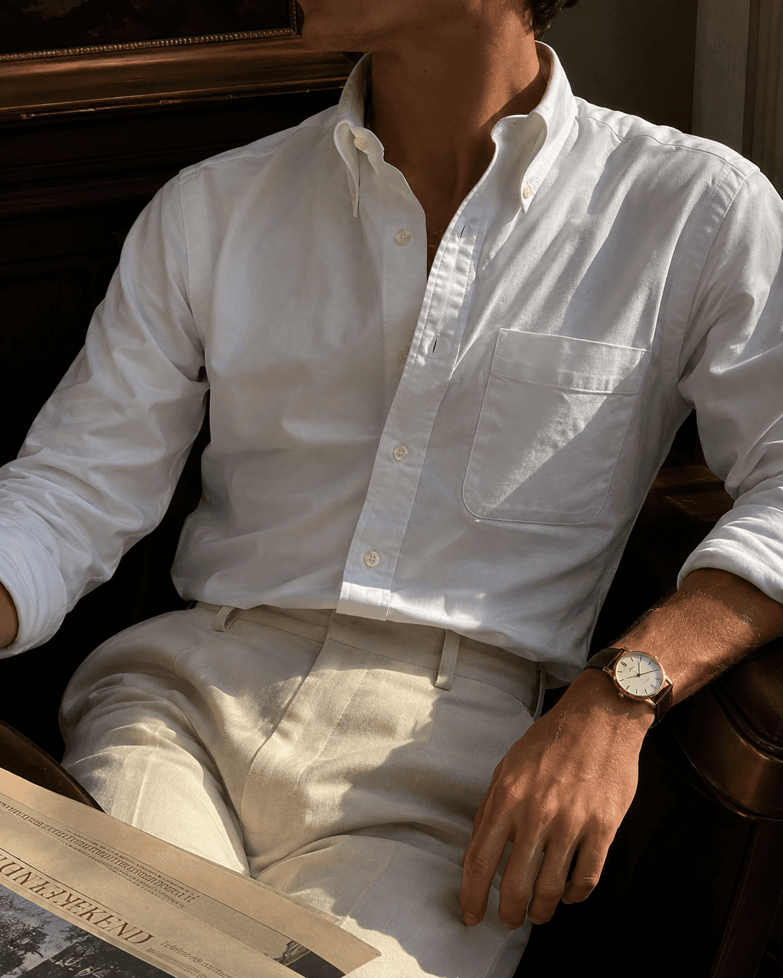 Midsection view of a man sitting in an armchair wearing men's classic old money style fashion, featuring a crisp white cotton oxford cloth button-down shirt with rolled sleeves, tucked into off-white pleated dress trousers, accessorized with a luxury gold wrist watch with a brown leather strap while holding a newspaper.