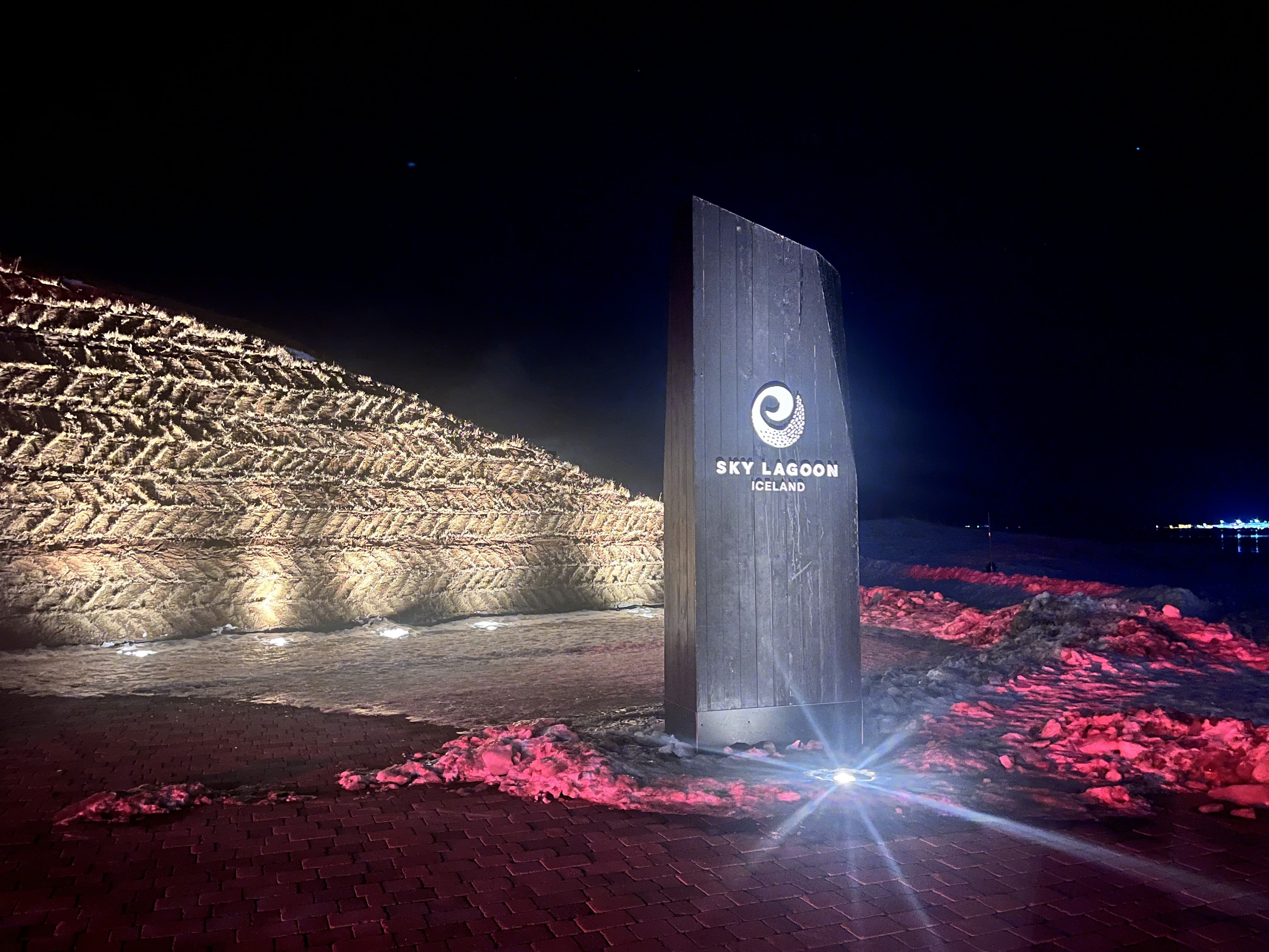 The entrance to a visitor center or exhibition with illuminated signage reading “Keflavík Airport” at night, surrounded by modern lighting and red ground lights.