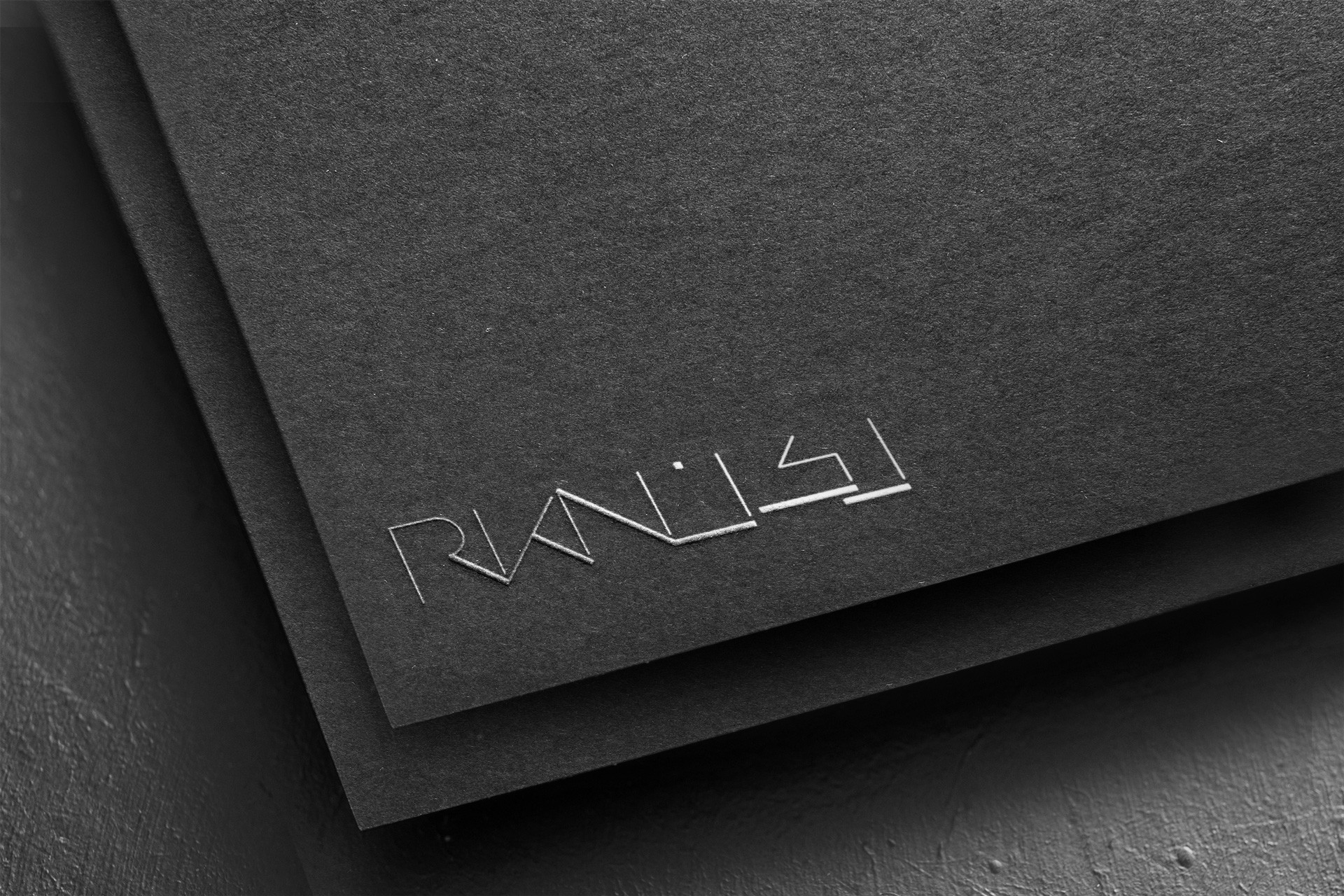 The RKN wordmark in silver foil, embossed on layered matte-black card, catching the light at an angle.