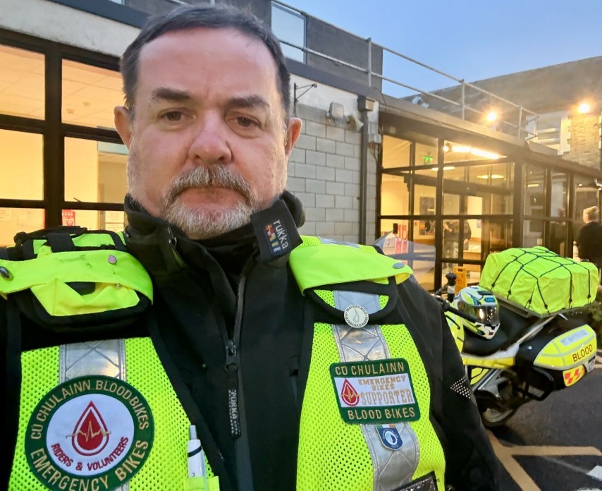 Smiling Blood Bikes volunteer, Andrew Ambrose, in motorcycle gear standing on a roadside with a castle in the background.