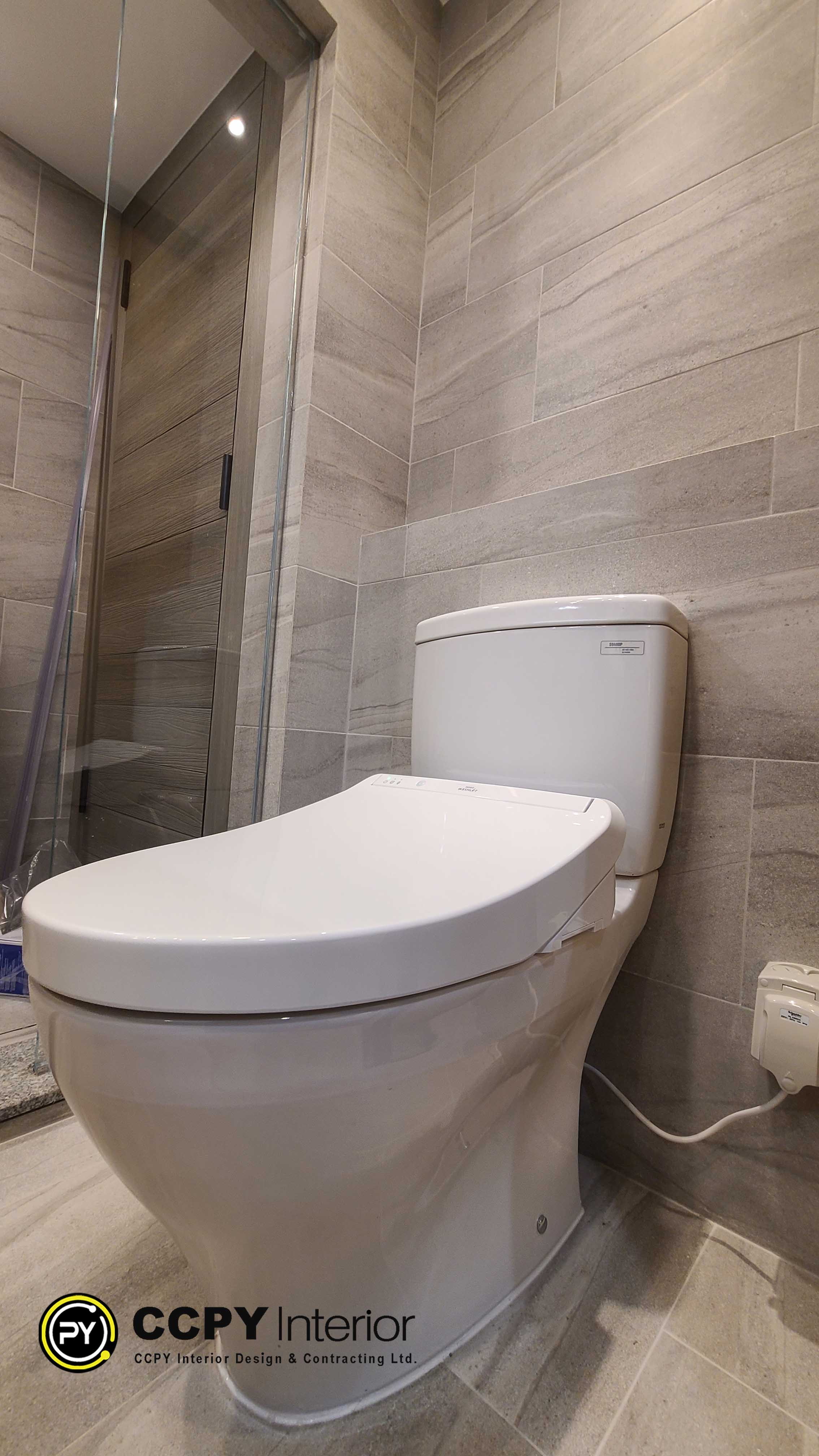 he selected region features a modern, close-coupled ceramic toilet equipped with an electronic bidet seat attachment, set against a backdrop of grey stone-look wall tiles.Visual Design FeaturesToilet Fixture: A white ceramic toilet with a classic rectangular tank and a curved, ergonomic bowl.Bidet Attachment: A sleek electronic bidet seat with a raised rear console housing the internal mechanics.Wall Tiling: Large-format ceramic or porcelain tiles featuring a matte, textured grey stone appearance with horizontal graining.Glass Enclosure: A partial view of a frameless glass shower panel or door visible on the left side.