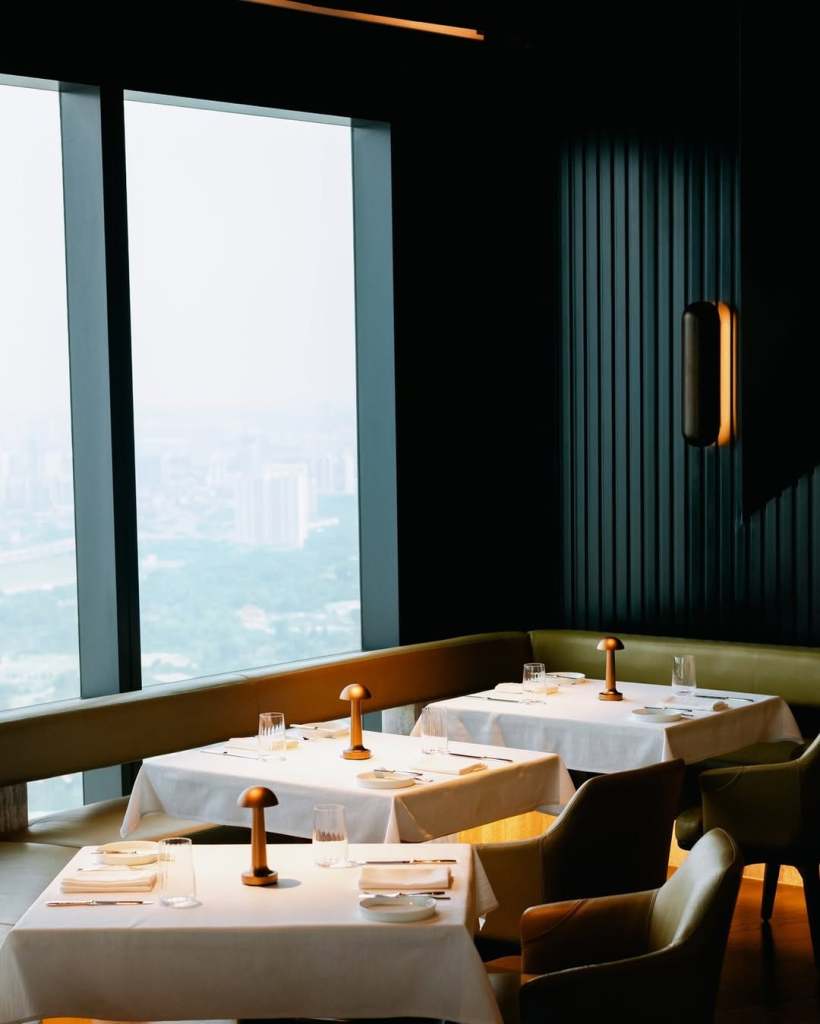 Elegant restaurant interior featuring white tablecloths, modern lighting, and panoramic city views through large windows.