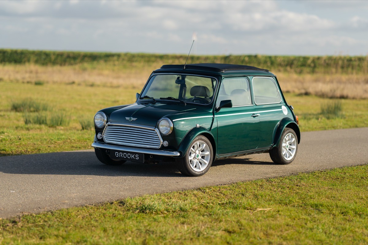 A vintage green car parked on a rural road with grassland in the background under a clear sky.